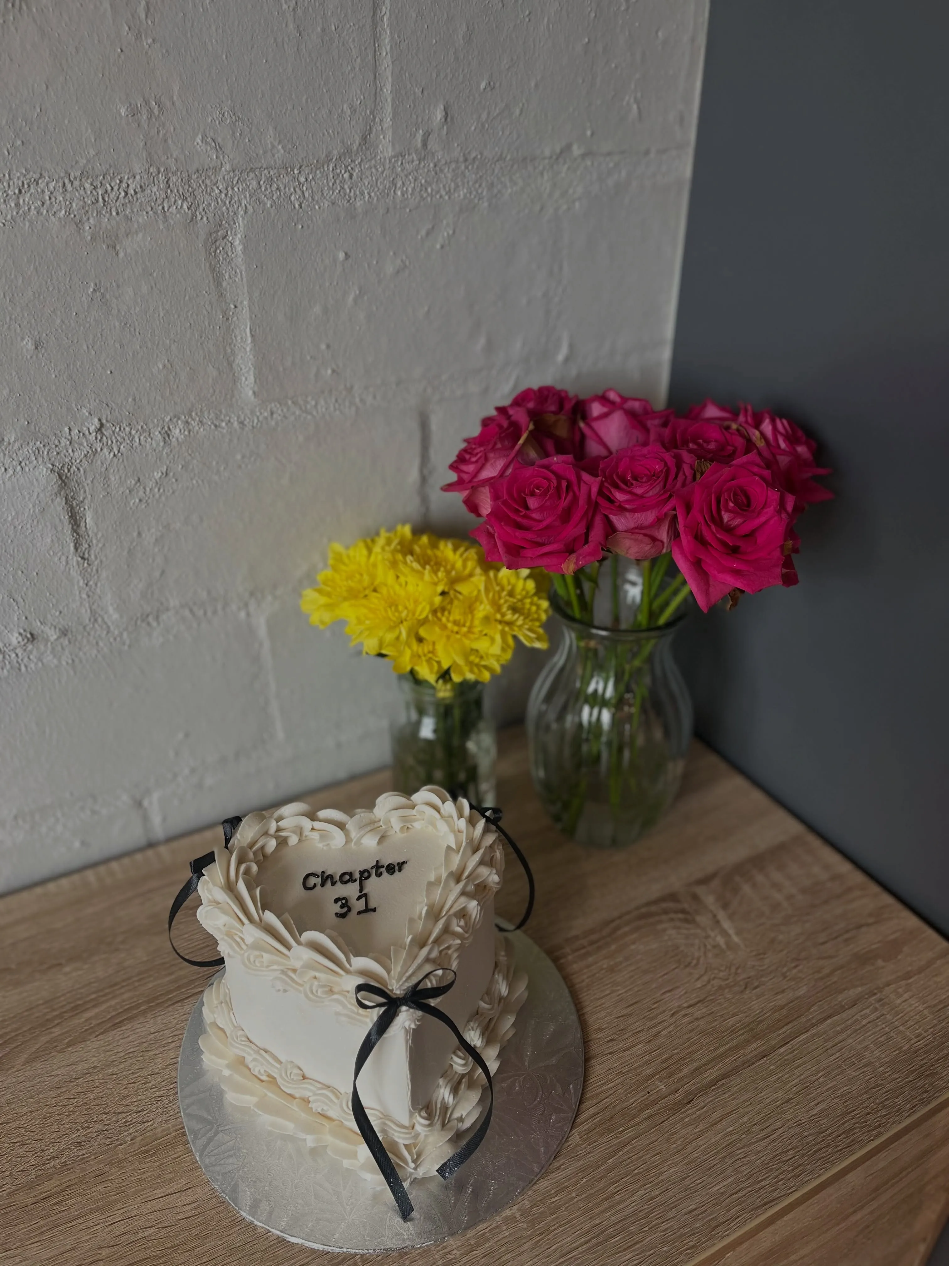 Cream heart cake with ruffles, a black bow and 'Chapter 31' piped on top, beside pink roses